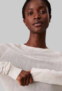 Maglione di un leggero beige a maglia con maniche lunghe e polsini a coste, caratterizzato da una superficie testurizzata e un collo a giro. Vista ravvicinata del collo e delle maniche.