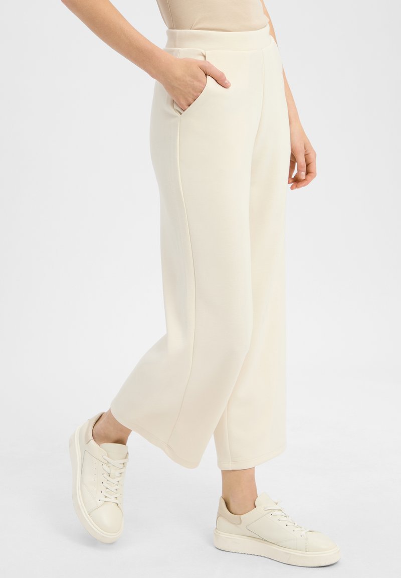 Crème brede broek gemaakt van zachte stof, met zijzakken en een gladde textuur, gecombineerd met lichtgekleurde veterschoenen.
