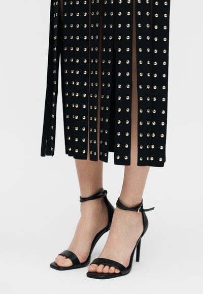 Μαύρα σανδάλια με ψηλό τακούνι και λουράκια στον αστράγαλο, φορεμένα με μαύρη φούστα που διαθέτει κάθετες εγκοπές και ασημένια καρφιά.