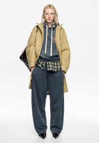 Persona rubia con un abrigo de lana beige oversized sobre una sudadera azul, camisa a cuadros verde, pantalones azules sueltos y zapatos negros de punta, de pie con las manos en los bolsillos.