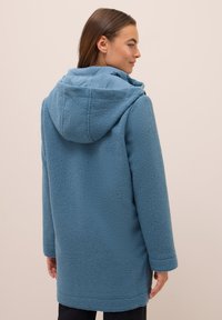 Manteau en polaire bleu doux avec capuche, présentant un extérieur texturé et des manches longues, porté sur un pantalon noir.