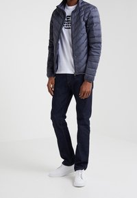 Veste matelassée grise avec col haut, associée à un t-shirt graphique blanc, un jean foncé et des baskets blanches. Fermeture éclair à l'avant et poches latérales.