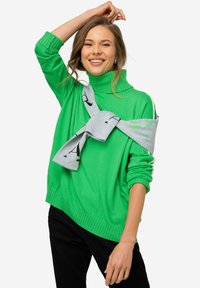 Maglione verde a collo alto con polsini a coste, indossato sopra un maglione grigio legato davanti. Pantaloni neri completano l'outfit.