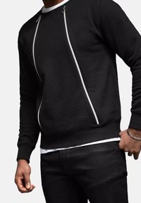 Sudadera negra con cremalleras blancas, puños y bajo acanalados. Presenta un diseño elegante con un acabado texturizado. Llevada sobre una camisa blanca.