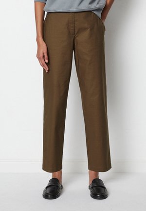 Persona con pantalones marrones de pierna recta con bolsillos laterales y mocasines negros sin cordones, de pie sobre un fondo blanco liso.