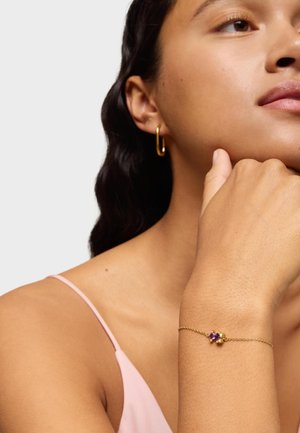 Pulsera de cadena de oro con piedras preciosas moradas. La modelo lleva pendientes de aro de oro y una blusa rosa, mostrando la pulsera en su muñeca.