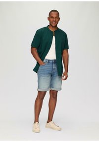 Groene kortekoolmouwen overhemd over een witte tanktop, gecombineerd met lichtblauwe denim shorts en beige sneakers. Eenvoudig en casual ontwerp.