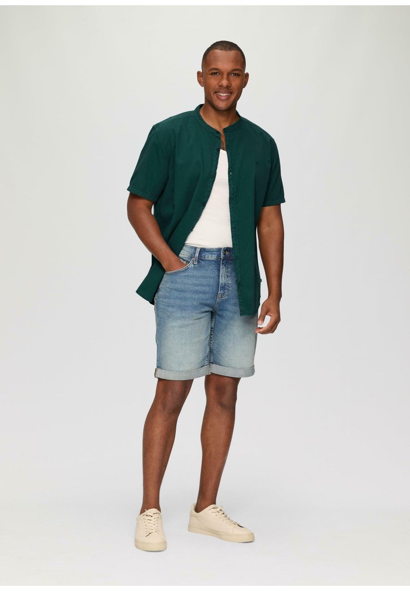 Groene kortekoolmouwen overhemd over een witte tanktop, gecombineerd met lichtblauwe denim shorts en beige sneakers. Eenvoudig en casual ontwerp.