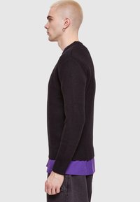 Pulover tricotat negru cu guler rotund, manșete și tiv ribbate, purtat de o persoană care are un tricou mov dedesubt. Vedere laterală.