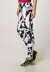 Mujer vistiendo leggings con un patrón abstracto en blanco y negro, zapatillas deportivas negras y verde neón, y una camiseta roja, de pie de lado.