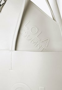 Biała skórzana torba z wytłoczonym napisem "LOLA CSDMNT" częściowo widoczna w większej białej skórzanej torbie.
