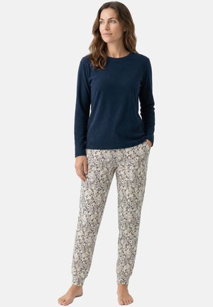 Vrouw staat blootsvoets, draagt een marineblauw shirt met lange mouwen en gebroken witte broek met bloemenpatroon, kijkt opzij tegen een effen achtergrond.