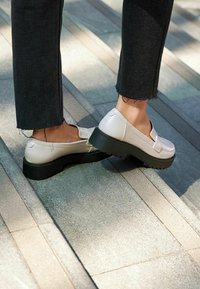 Zapatos de plataforma de color crema con suela negra de goma, material suave similar al cuero y diseño sin cordones. Combinados con pantalones negros desgastados.