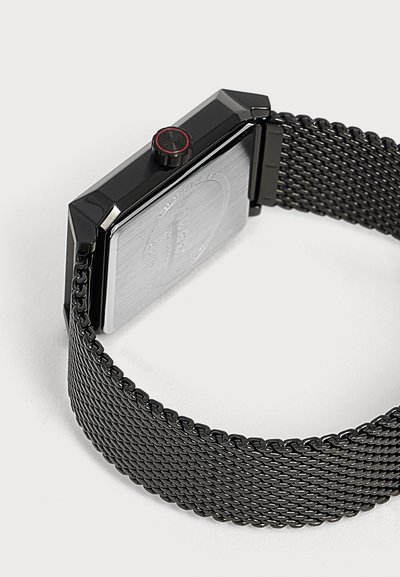 Bracelet de montre en maille métallique noire avec un fond de boîtier rectangulaire et une couronne latérale ronde avec un accent d'anneau rouge sur fond gris.