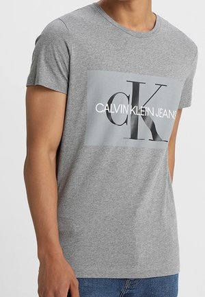 Homme portant un t-shirt gris à manches courtes avec un grand logo rectangulaire Calvin Klein Jeans en noir et blanc sur la poitrine.