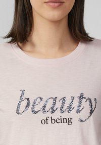 T-shirt rose clair avec le texte noir "de l'être" et le mot "beauté" en script pailleté dans des nuances d'argent et de bleu. Tissu doux avec un col rond.