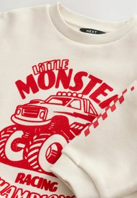 Licht beige sweatshirt met rode graphic van een monstertruck en de tekst "Little Monster Racing Champions." Ribbelkraag en -manchetten.