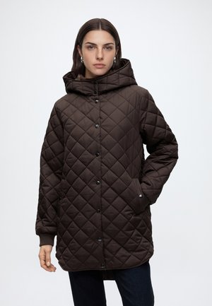 Femme portant une veste matelassée à capuche marron foncé avec des boutons-pression et les mains dans les poches, sur un fond clair uni.