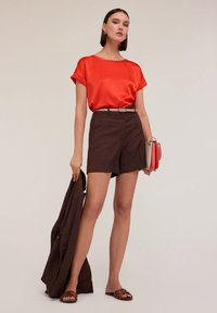 Haut rouge en satin à manches courtes, short marron ajusté, ceinture marron et veste marron. Les accessoires comprennent un sac à main multicolore et des sandales marron.