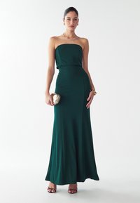 Naine kannab rihmata tumerohelist põrandani ulatuvat kleiti, hoides kuldset kotikest, kaunistatud kuldsete kõrvarõngaste, käevõru ja kaelakeega, seistes.