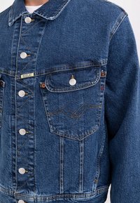 Blå denimjacka med en klassisk krage, framknäppning, två bröstfickor och dekorativ sömnad på vänster ficka.