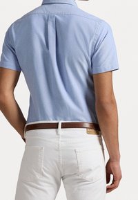 Muž v světle modré košili s krátkým rukávem zastrčené do bílých kalhot s hnědým koženým pásem, zobrazený zezadu.