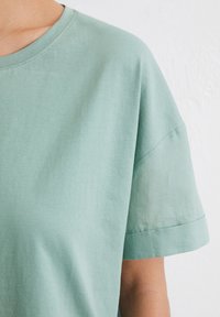 Hellgrünes T-Shirt mit kurzen Ärmeln, Rundhalsausschnitt, weicher Baumwollstoff, lockere Passform, sichtbare Nahtdetails an Ärmeln und Ausschnitt.