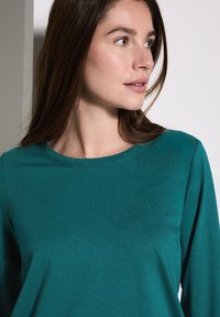 Teal langetermijn shirt van zachte stof, met een ronde hals en een gladde textuur. Het shirt heeft een losse pasvorm en een minimaal ontwerp.