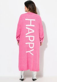 Cardigan lungo rosa realizzato in materiale a maglia, con la parola "HAPPY" scritta in lettere bianche sul retro. Le maniche sono sagomate in modo morbido.