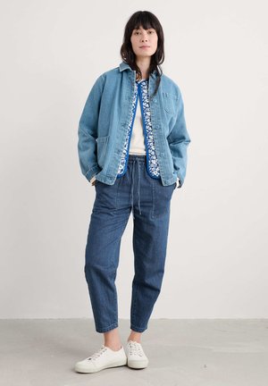 Lichte blauwe spijkerjack, witte gestreepte shirt en donkerblauwe slim fit broek, gecombineerd met witte sneakers. Heeft een ontspannen pasvorm en zakken.