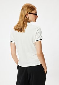 Witte polo met zwarte rand, korte mouwen en een getailleerd ontwerp. De stof heeft een gestructureerde, geribbelde afwerking. Zwarte broek zichtbaar.