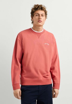 Jonge man met krullend haar, die een koraalkleurige sweatshirt met het "Pepe Jeans"-logo en een donkere broek draagt, staand tegen een effen lichte achtergrond.