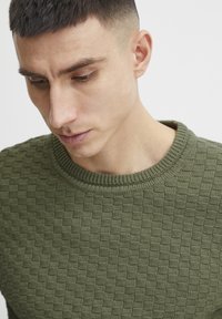 Groene gebreide trui met een getextureerd mandweefpatroon, ronde halslijn en geribbelde randen. De stof lijkt zacht en dik te zijn.