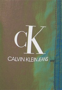 Nahaufnahme eines grünen Stoffes mit dem weißen Logo "ck CALVIN KLEIN JEANS" und sichtbaren Nähten auf der rechten Seite.
