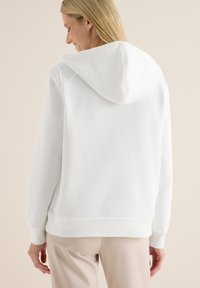 Felpa bianca con cappuccio, dotata di polsini e orlo a costine, caratterizzata da una texture liscia e una vestibilità comoda. Cuciture visibili lungo le spalle.