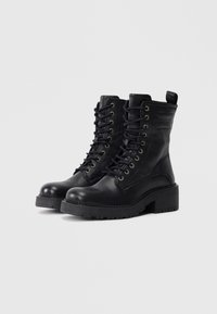 Botas de tobillo de cuero negro con diseño de cordones, ojales dorados, punta redonda y un tacón grueso con suela texturizada.