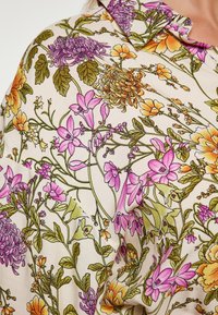 Blusa floreale con sfondo crema pastello, caratterizzata da grandi fiori rosa e arancioni, foglie verdi e un colletto.