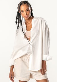 White oversized cotton shirt, buttoned, with a relaxed fit and rolled-up sleeves, paired with beige linen shorts. Smooth texture, no patterns.