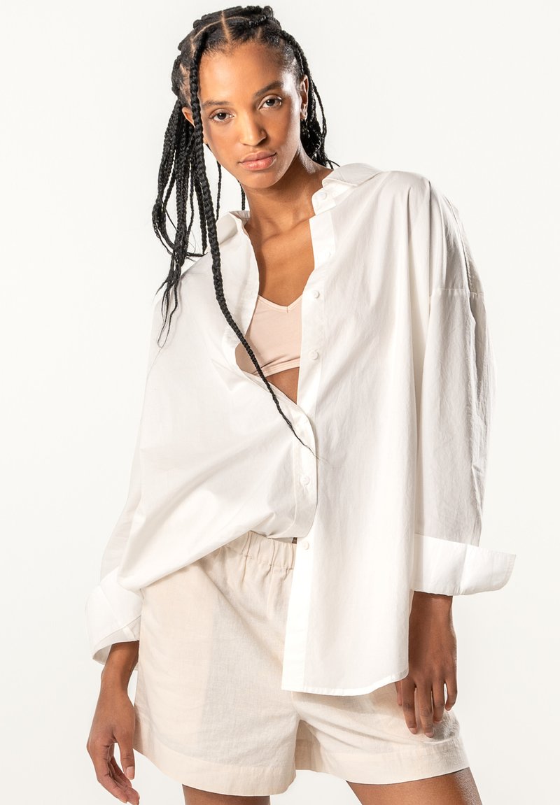 White oversized cotton shirt, buttoned, with a relaxed fit and rolled-up sleeves, paired with beige linen shorts. Smooth texture, no patterns.