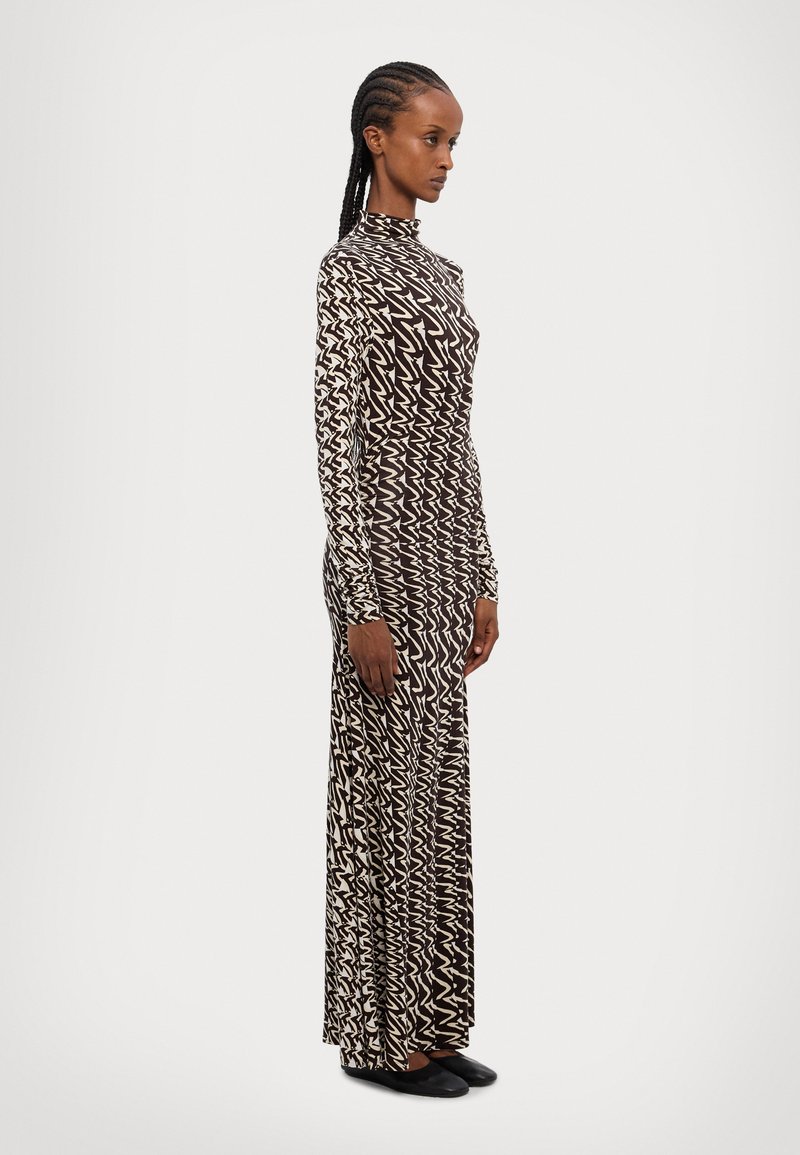 Robe à manches longues, avec un motif zigzag noir et blanc, un col haut, une silhouette ajustée et un tissu lisse, accompagnée de chaussures plates noires.