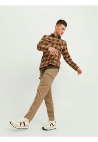Camicia a quadri marrone, pantaloni cargo beige e sneakers bianche con dettagli rossi. L'outfit presenta un design informale e una vestibilità rilassata.