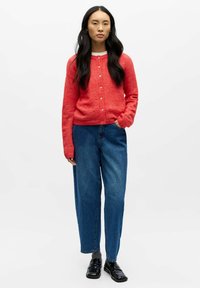 Sarkans vilnas kardigans ar apaļu kakla izgriezumu un pogu aizdari, kopā ar vaļīgām zilām džinsa biksēm un melniem spīdīgiem apaviem. Vienkāršs balts fons.