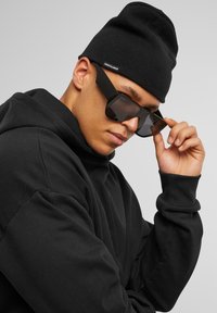 Bonnet noir et hoodie noir surdimensionné, associés à de grandes lunettes de soleil noires à verres foncés, montrant un profil rapproché.
