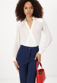 Balta permatoma palaidinė su V formos iškirpte, ilgomis rankovėmis ir kontrastingais manžetais, derinama su tamsiai mėlynomis pritaikytomis kelnėmis ir raudona rankine.