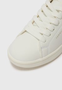Weiße Leder Turnschuhe mit flachen Schnürsenkeln, abgerundetem Zehenbereich und subtilen, tonalen Nähten. Mit einem goldenen Logo an der Seite. Glatte Textur.