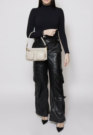 Femme portant un col roulé noir et un pantalon cargo large en cuir noir, tenant un sac à main beige avec des poches avant, et chaussée de talons noirs cloutés.