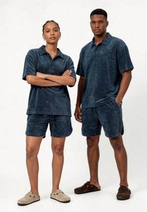 Dva modeli nosita ujemna temno modra polo majica iz frote in kratke hlače, stoje pred enostavno svetlo ozadje.