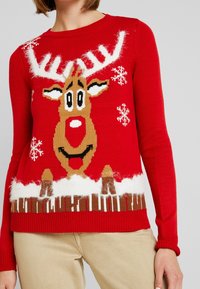 Punainen neulottu villapaita, jossa on valko- ja ruskeasävyinen porokuvio, lumihiutalekuvioita sekä pehmeää yksityiskohtaa pääntiellä ja helmassa.