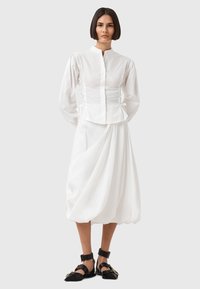 Biała koszula z wysokim kołnierzem, bufiastymi rękawami i bocznymi sznurowaniami. W połączeniu z obszerną białą spódnicą i czarnymi sandałami na paski z złotymi akcentami.