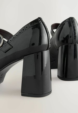 Schwarze lacklederne High-Heels mit dicken, quadratischen Absätzen, Brogue-Details und einem Schnallenriemen über dem Mittelfuß. Glatte, glänzende Oberfläche.